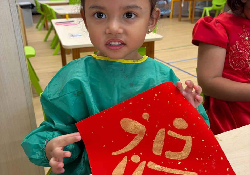 Child in green smock holds red paper with gold calligraphy, standing in a classroom with green chairs.